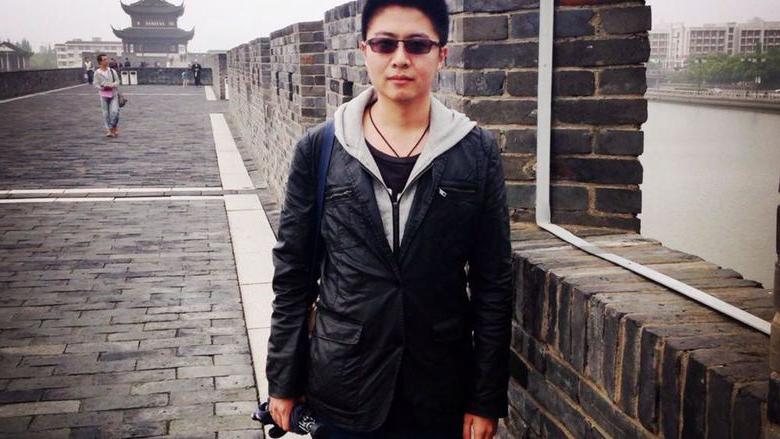 A person in sunglasses and a light jacket poses in front of a brick wall on a gray day. Buildings with traditional Chinese and with modern architecture can be seen in the background. 